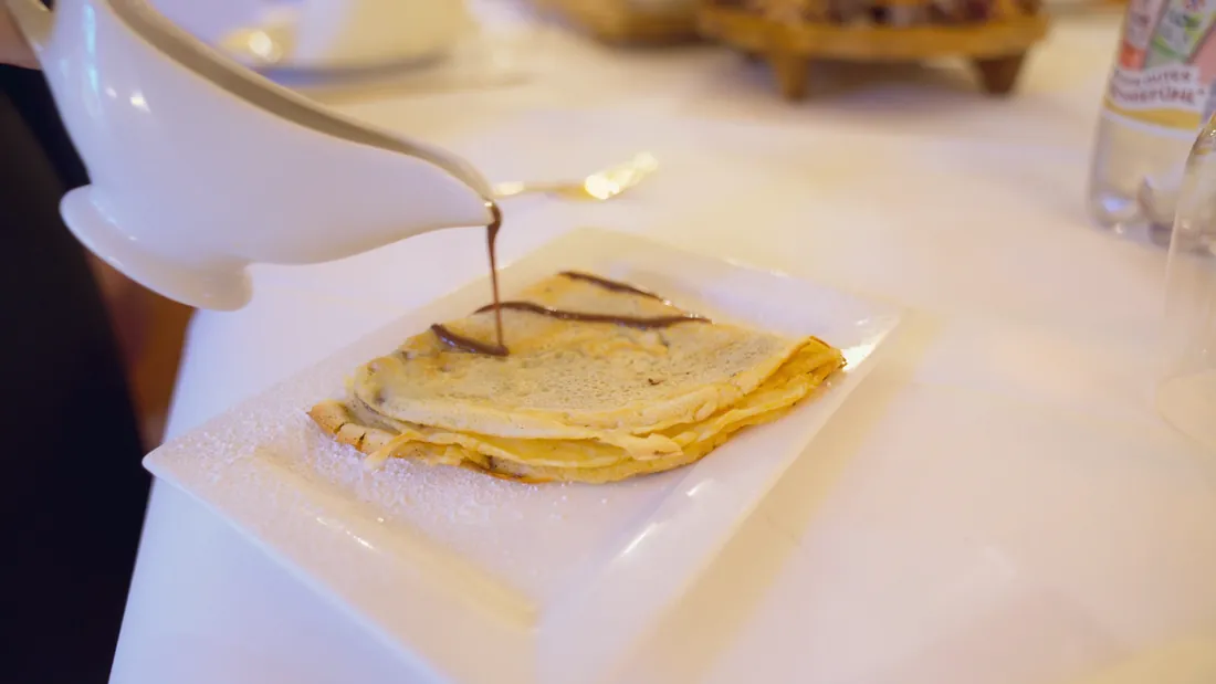 Crêpes auf einem weißen Teller, Schokoladensauce wird darüber gegossen